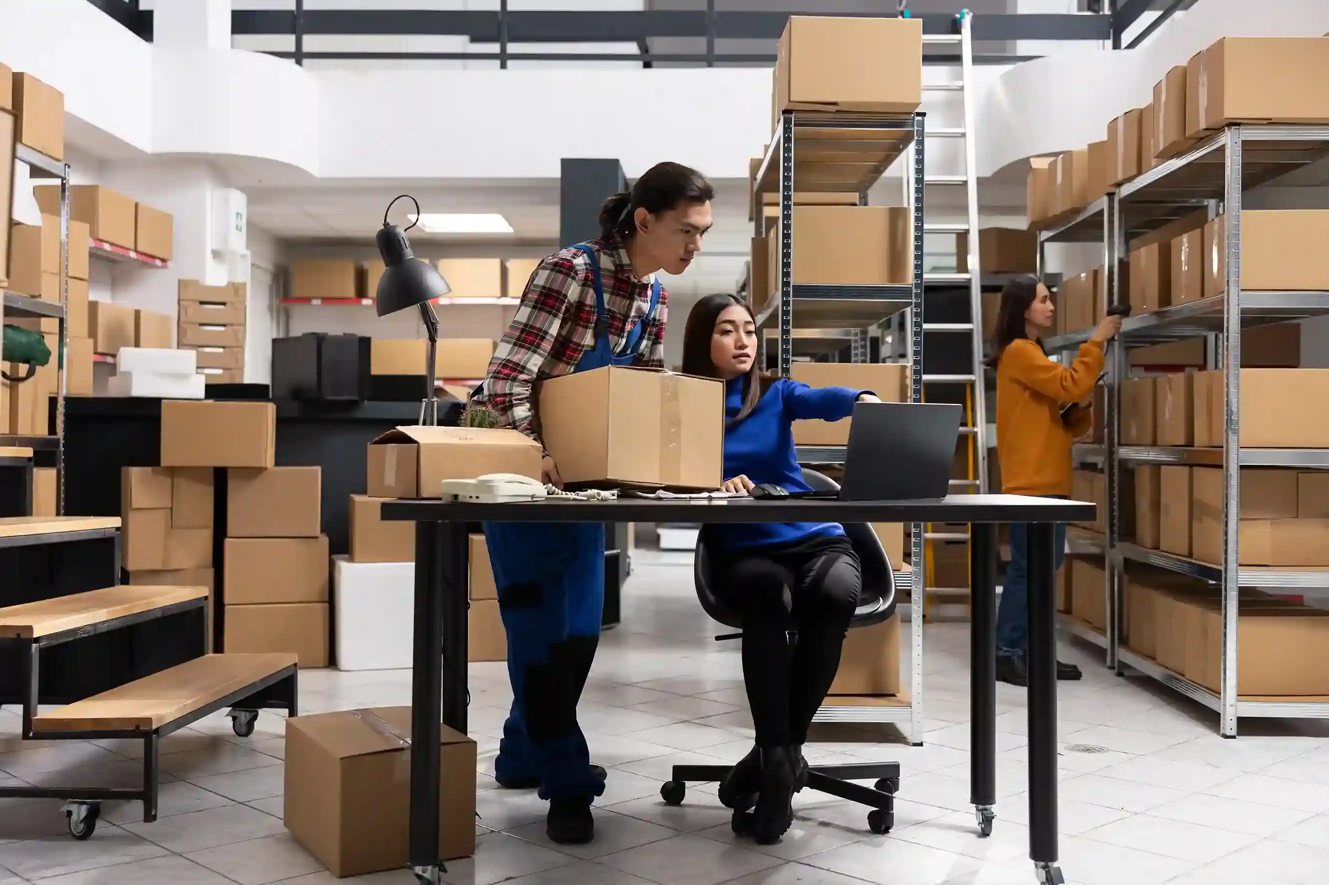Small business team managing inventory and packaging boxes in a warehouse workspace using a laptop for operations.